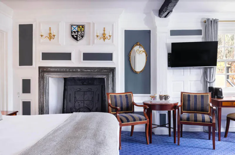 Elegant Tudor-style bedroom at Karma Salford Hall with fireplace, armchairs, table, TV, and crest wall decor.