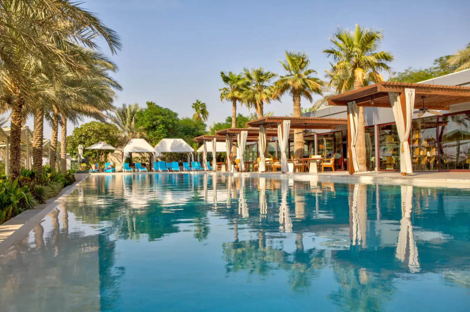 Luxury pool at Melia Desert Palm Dubai resort, surrounded by palm trees and wooden cabanas under blue sky.