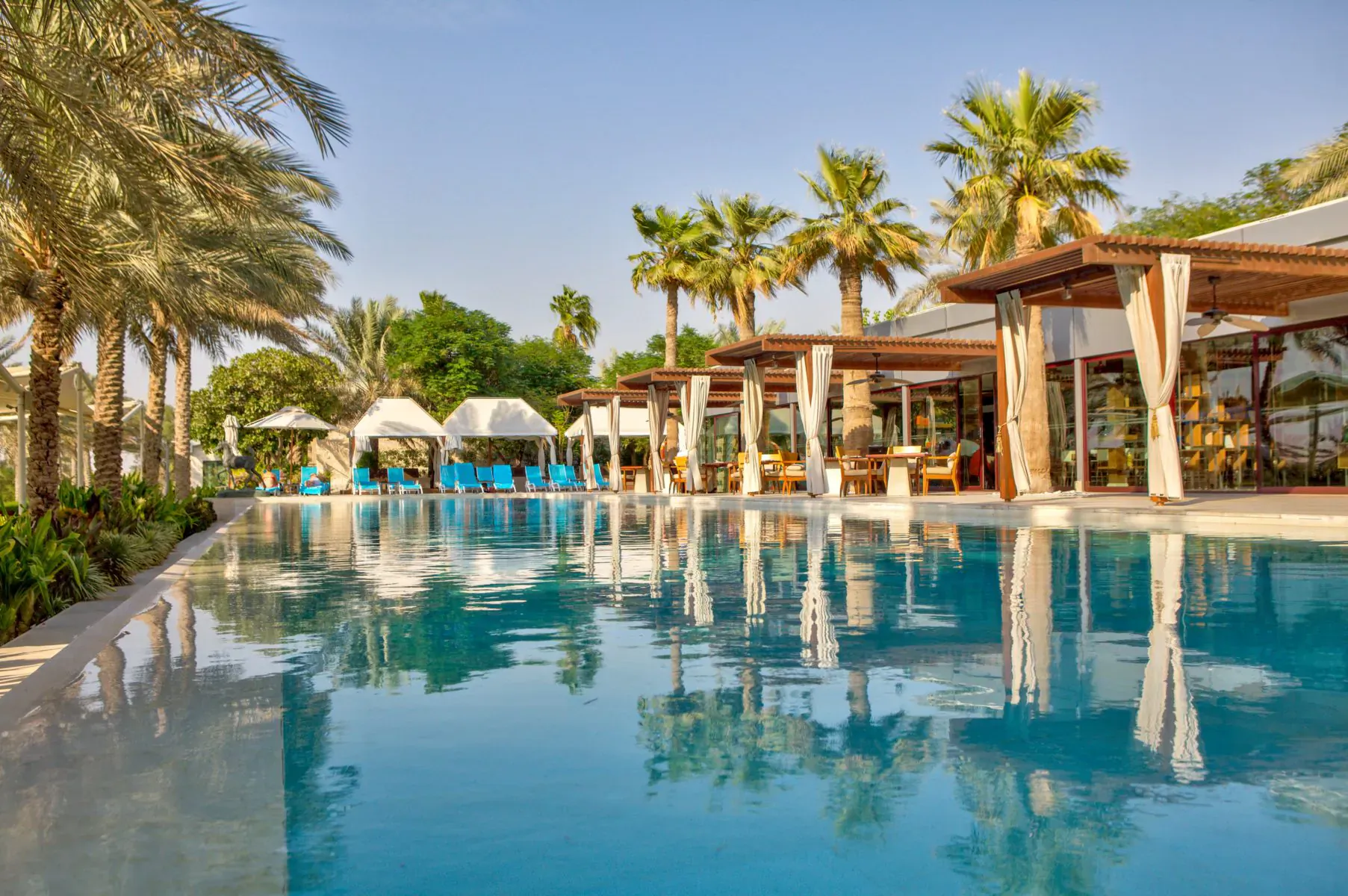 Luxury pool at Melia Desert Palm Dubai resort, surrounded by palm trees and wooden cabanas under blue sky.
