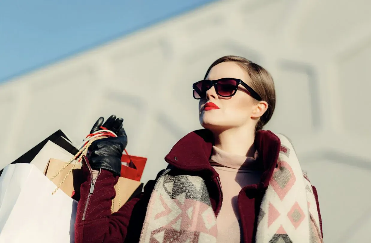 Stylish woman in sunglasses, red lipstick, gloves, and patterned scarf holding luxury shopping bags outdoors