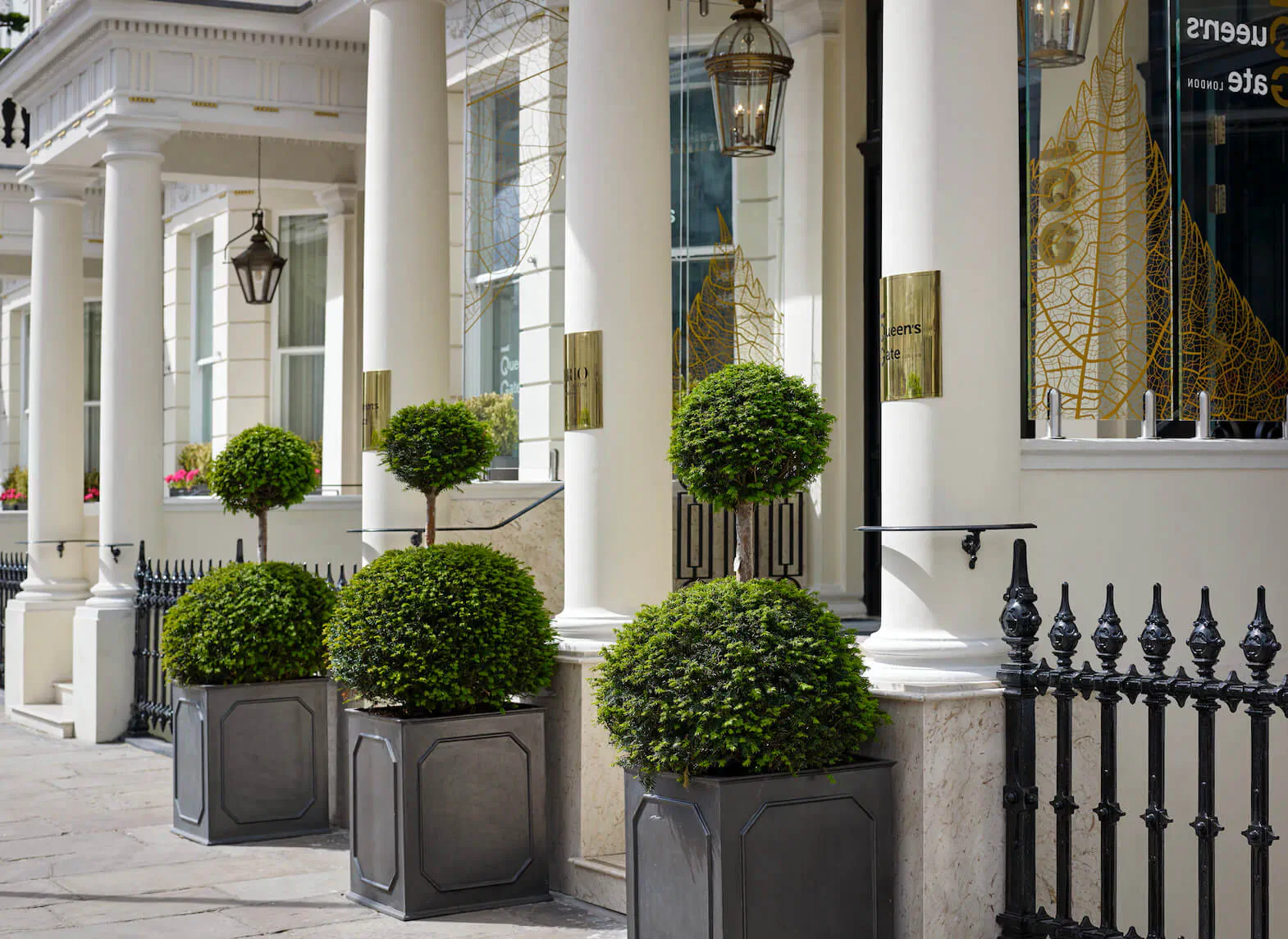 Elegant white-columned townhouse entrance in London with lanterns, topiary boxwoods in gray pots, and black railing.