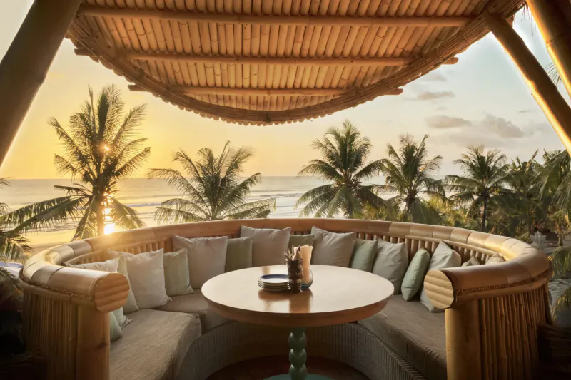 Sunset view of Azul Beach Club Bali: circular bamboo lounge with cushions, low table, tropical palms, and ocean horizon.