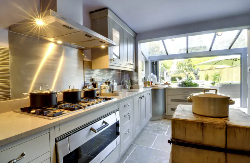 Modern luxury kitchen in cottage with white cabinets, gas stove, pots on counters, and garden view through glass doors.