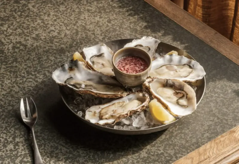 Oysters on ice in a round dish with lemon wedges and red sauce, spoon beside, on green stone table