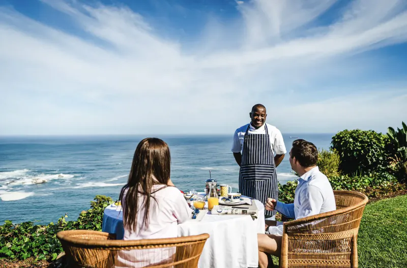 Outdoor breakfast table overlooking Atlantic ocean in Cape Town, smiling Black waiter in apron serves smiling white couple, sunny sky.