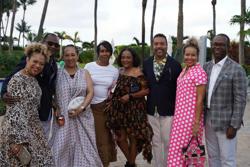 Group of six smiling Black adults in elegant attire posing outdoors among palm trees at Italian affair event.