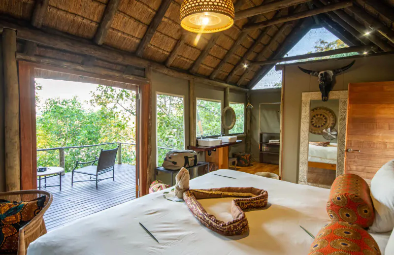 Luxurious thatched Botswana lodge room with king bed, coiled snake decor, orange pillows, and balcony view of Tsodilo Hills forest.