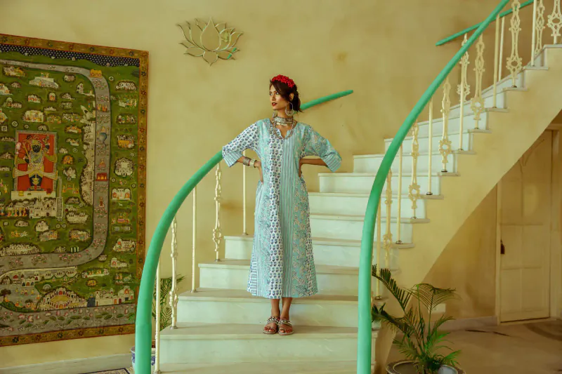 Woman in light blue embroidered maxi dress stands on white stairs with green railing, beside vibrant tapestry in gallery
