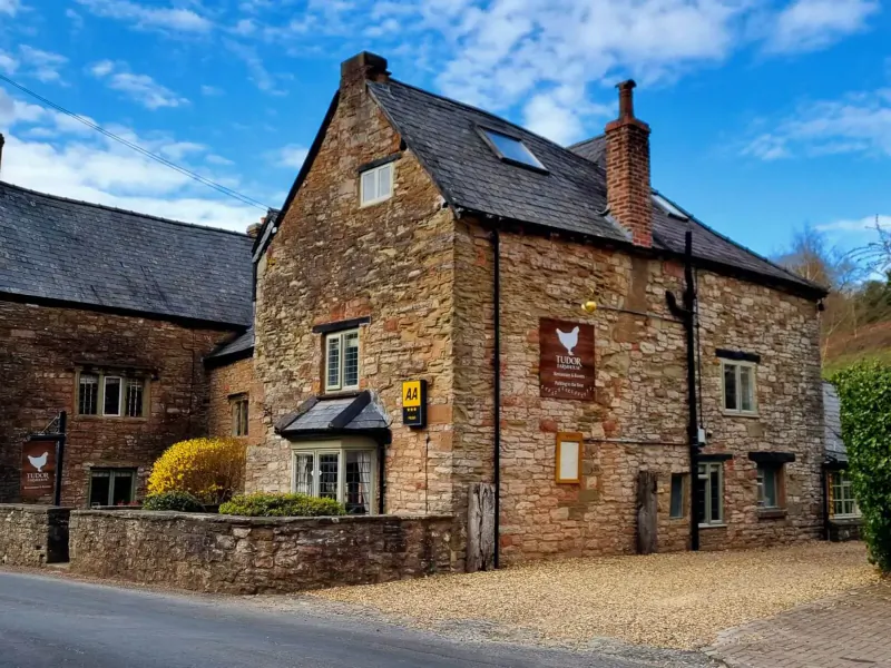 Tudor Farmhouse boutique hotel, stone building with golden hen sign, in Forest of Dean countryside under blue sky.