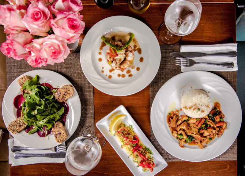Elegant restaurant table with pink roses, plates of shrimp dish, beet salad, rice with prawns, lemon salsa, wine glasses.