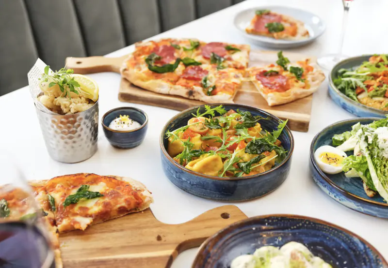 Assortment of pizzas with salami and greens, fries, Caesar salad, and pasta on a restaurant table