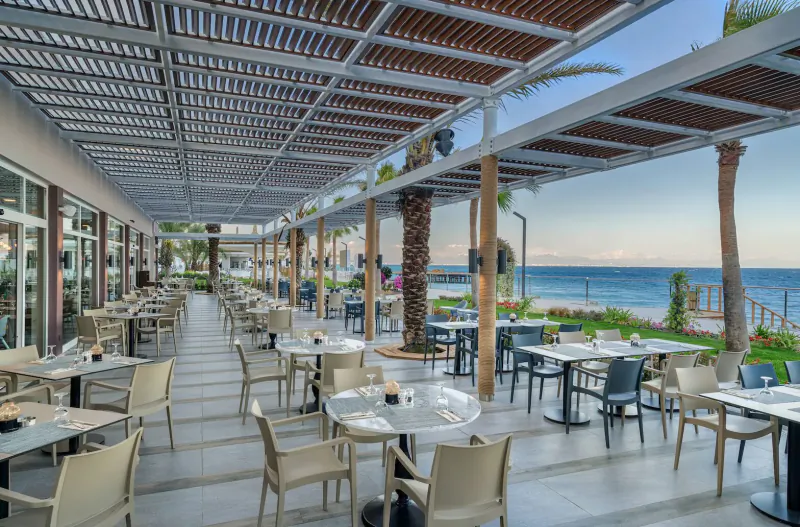 Outdoor terrace restaurant at Corendon Playa Kemer with tables, chairs, palms, and sea view at sunset.