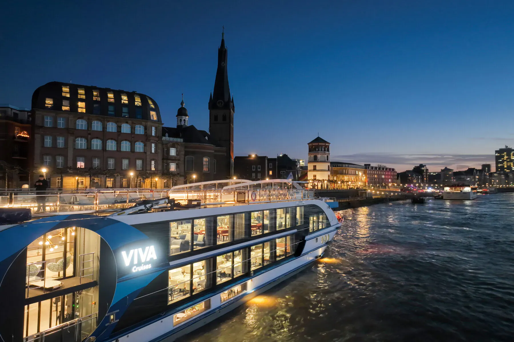 Viva Cruises luxury riverboat on Dutch waterway at dusk, with historic church tower and lit buildings.