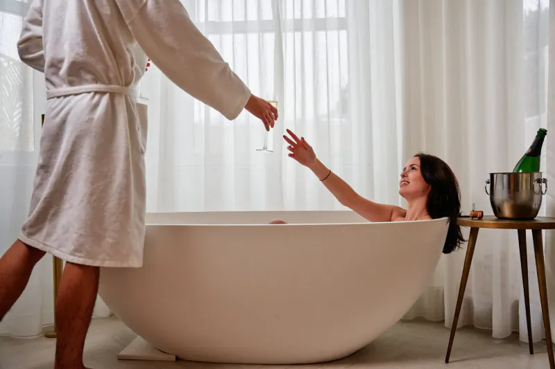 Man in white robe hands champagne flute to smiling woman relaxing in large white bathtub, bottle in ice bucket nearby, sheer curtains.