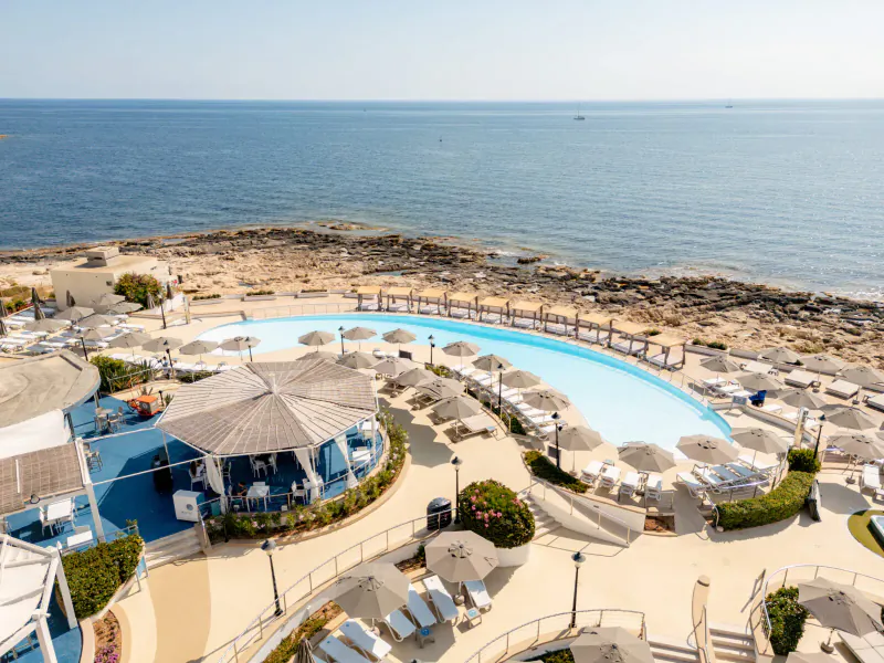 Aerial view of Hilton Malta's infinity pool, sun loungers, beach bar, and Mediterranean Sea.