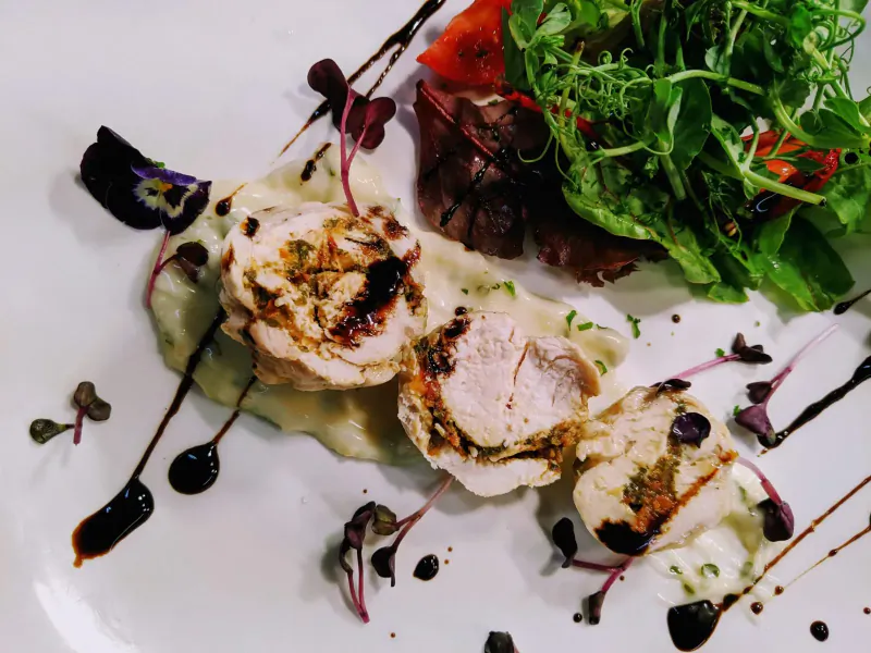 Three grilled chicken roulades on white plate with creamy mash, balsamic drizzle, red tomatoes, and microgreens