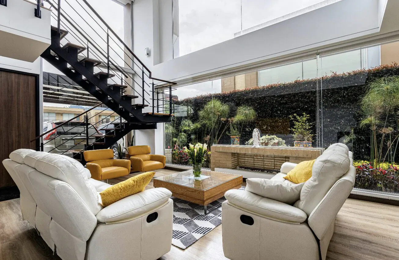 Bright modern atrium living area with white leather armchairs, yellow cushions, coffee table with flowers, black metal stairs, and lush green plants.
