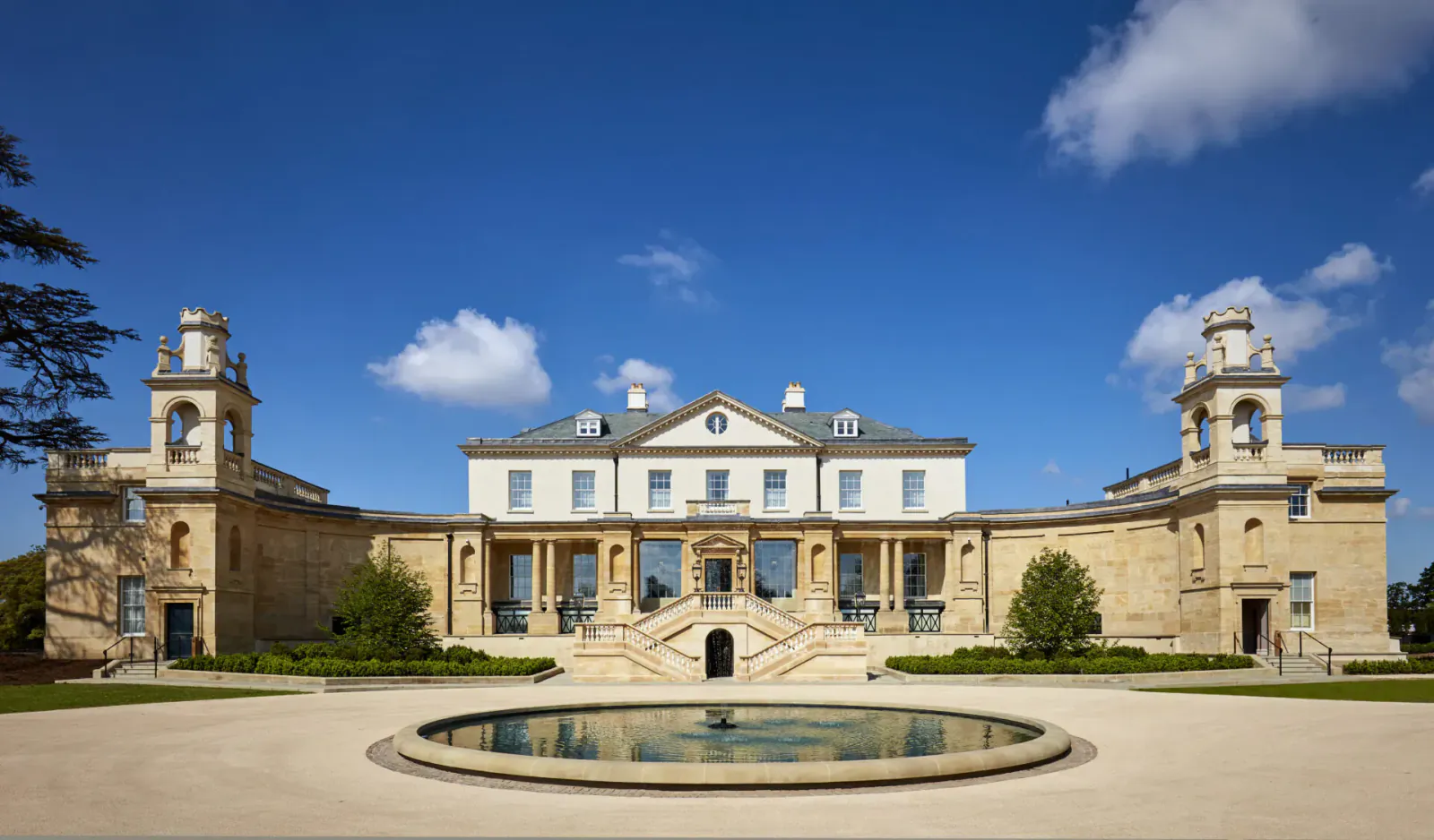 The Langley Hotel, a grand 5-star luxury mansion in Buckinghamshire with clock towers, steps, fountain, and gardens under blue sky.