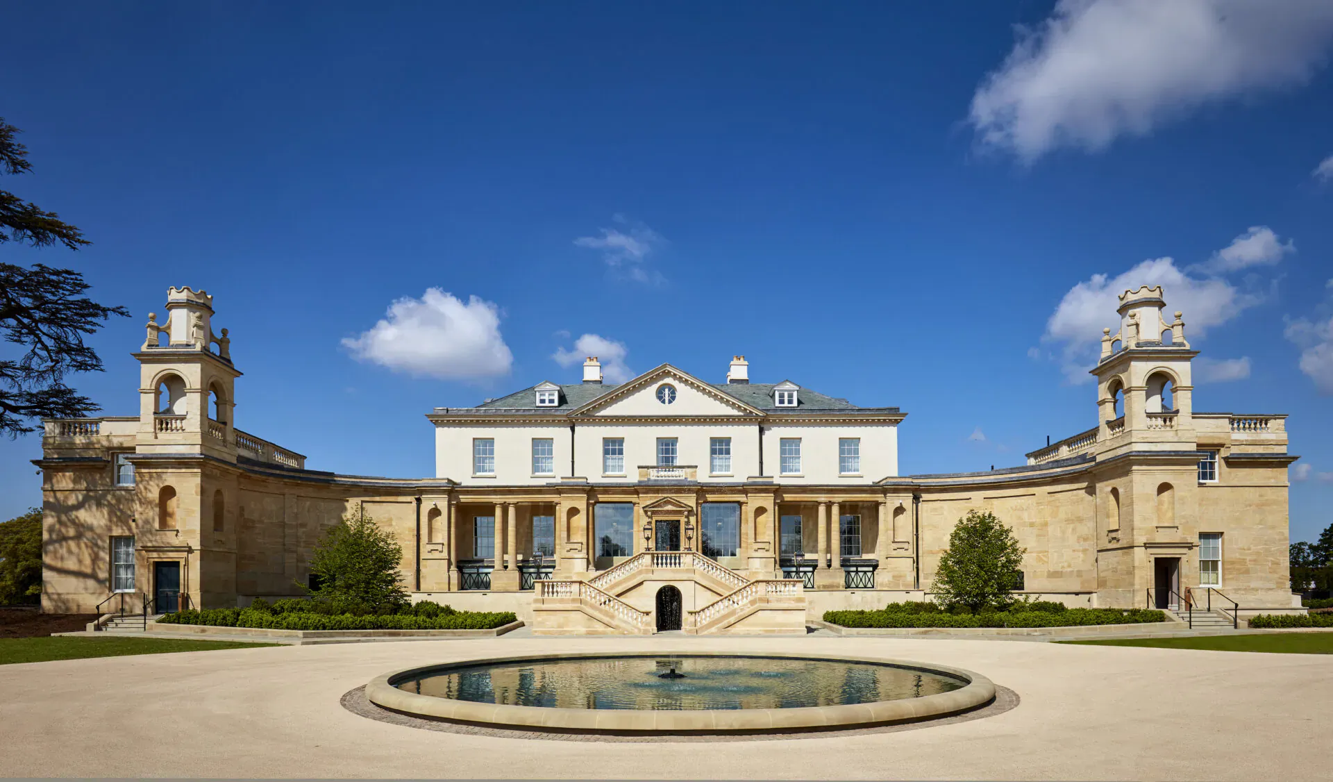 The Langley Hotel, a grand 5-star luxury mansion in Buckinghamshire with clock towers, steps, fountain, and gardens under blue sky.