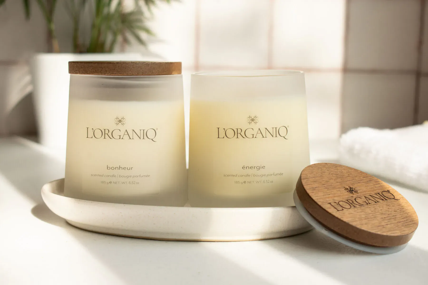 Two Lorganiq candles 'Sunset' and 'Sunrise' on white plate with wooden lids, on tiled surface with plant and towel.