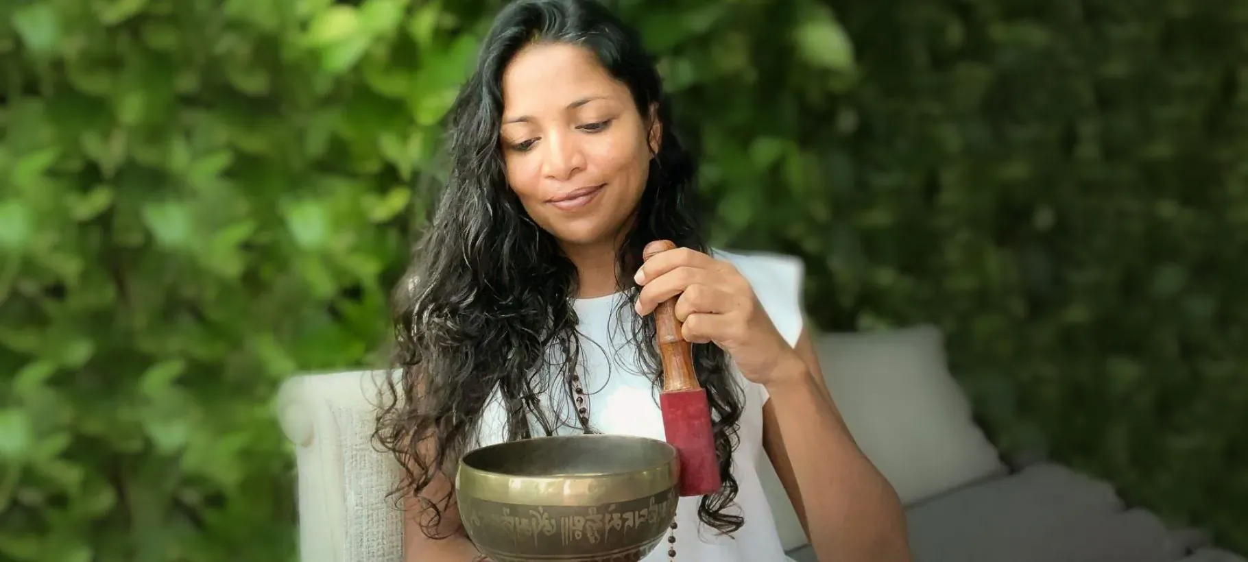 Smiling woman with long dark hair holds mallet to singing bowl, seated outdoors amid green foliage at wellness resort