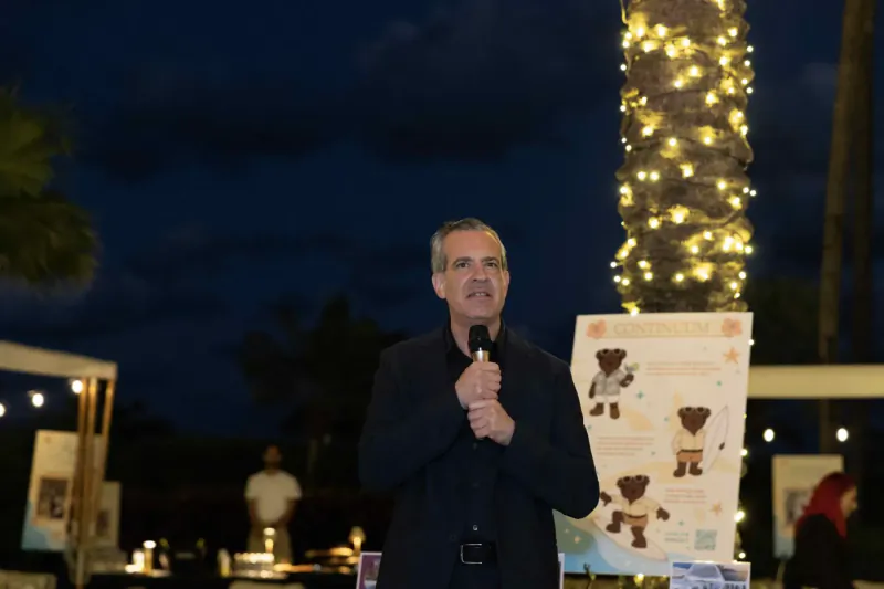 Man in dark suit speaking into microphone at nighttime outdoor event beside teddy bear poster and lit palm tree