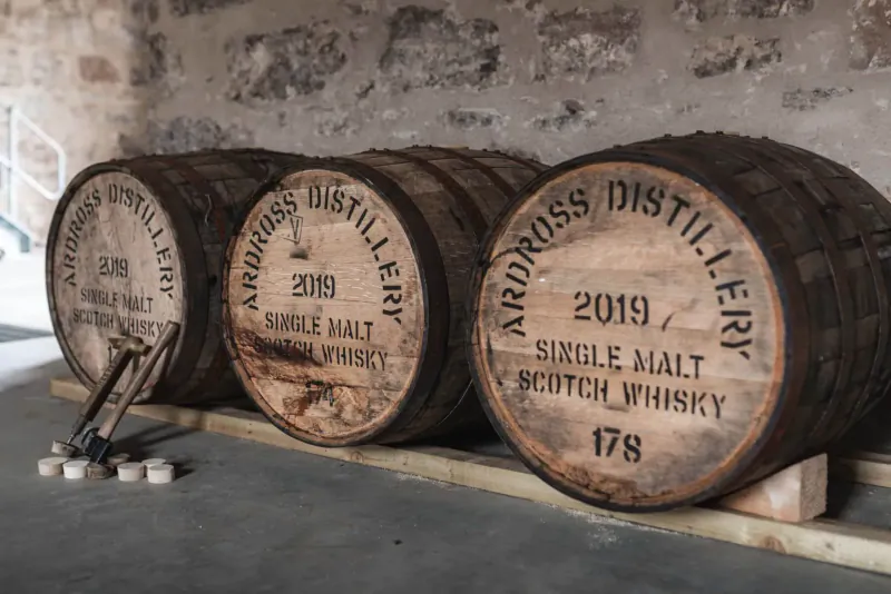 Three Ardross Distillery single malt whisky barrels labeled 2019 and 178, in stone-walled warehouse with barrel tools nearby.