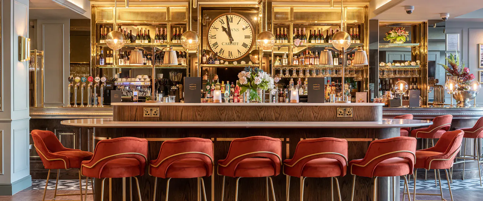 Elegant bar at Browns Cardiff with wooden counter, large gold clock, bottles, flowers, and red swivel stools.