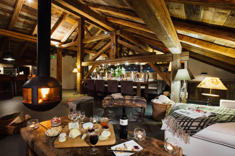 Cozy Alpine chalet interior with wooden beams, roaring hanging fireplace, cheese and wine board on table, plush seating.