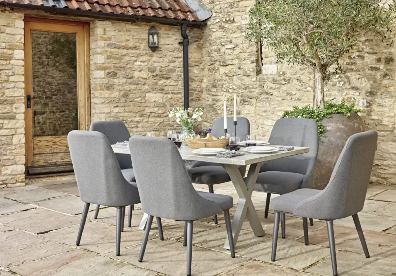 Grey upholstered 8-seat X-frame outdoor dining set on stone patio by stone wall with olive tree, lantern, flowers.