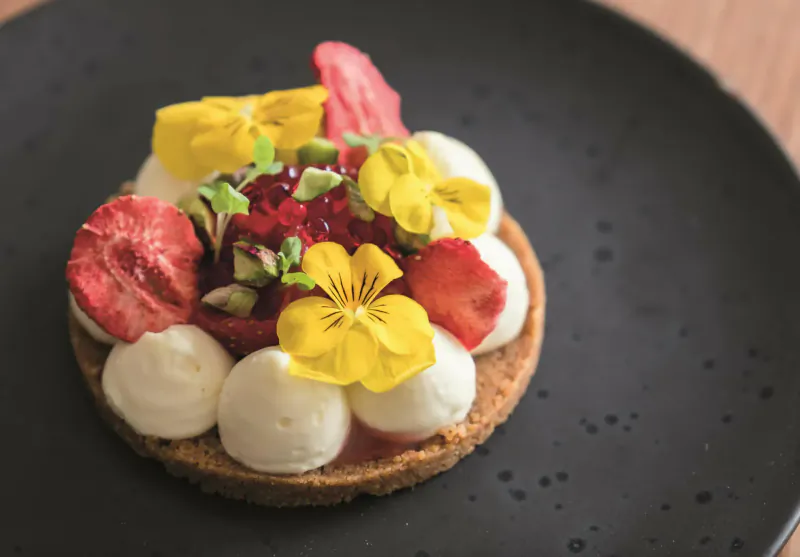 Strawberry tart topped with whipped cream, edible yellow viola flowers, and red berries on black plate