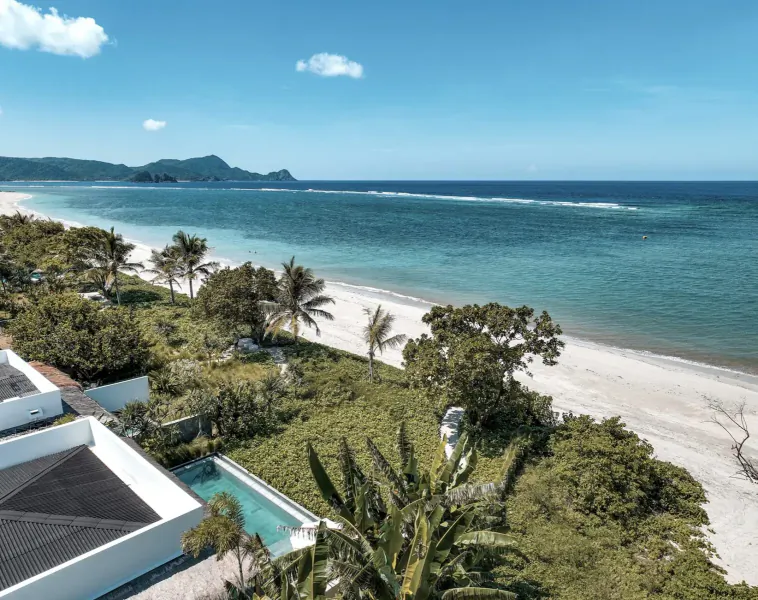 Aerial view of Amber Lombok Beach Resort: modern white villa with pool, lush gardens, palm trees, sandy beach, and ocean with mountains.