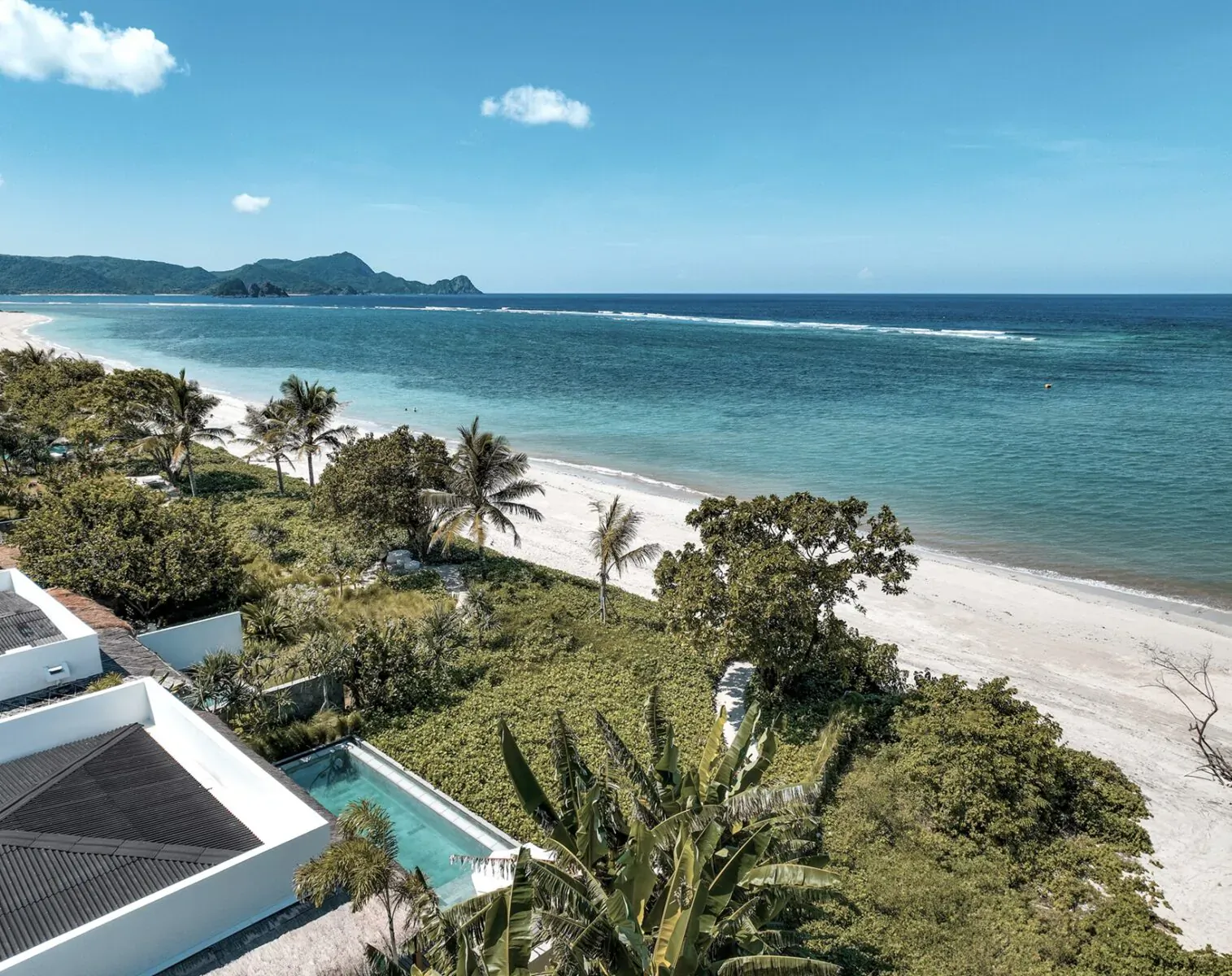 Aerial view of Amber Lombok Beach Resort: modern white villa with pool, lush gardens, palm trees, sandy beach, and ocean with mountains.