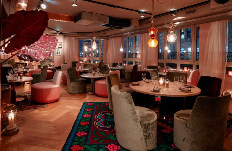 Cozy 25Hours Paris restaurant interior at night with velvet chairs, round tables, pendant lights, large windows, and patterned rug.