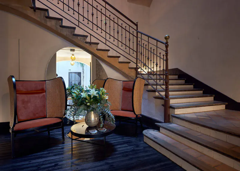 Elegant hotel lobby with curved red armchairs, floral vase on round table, ornate wooden staircase, and arched doorway.