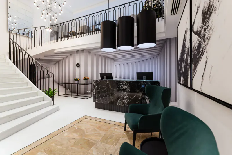 Elegant Embassy Valletta Hotel lobby with marble reception desk, green armchairs, curved white staircase, chandeliers, and striped walls.