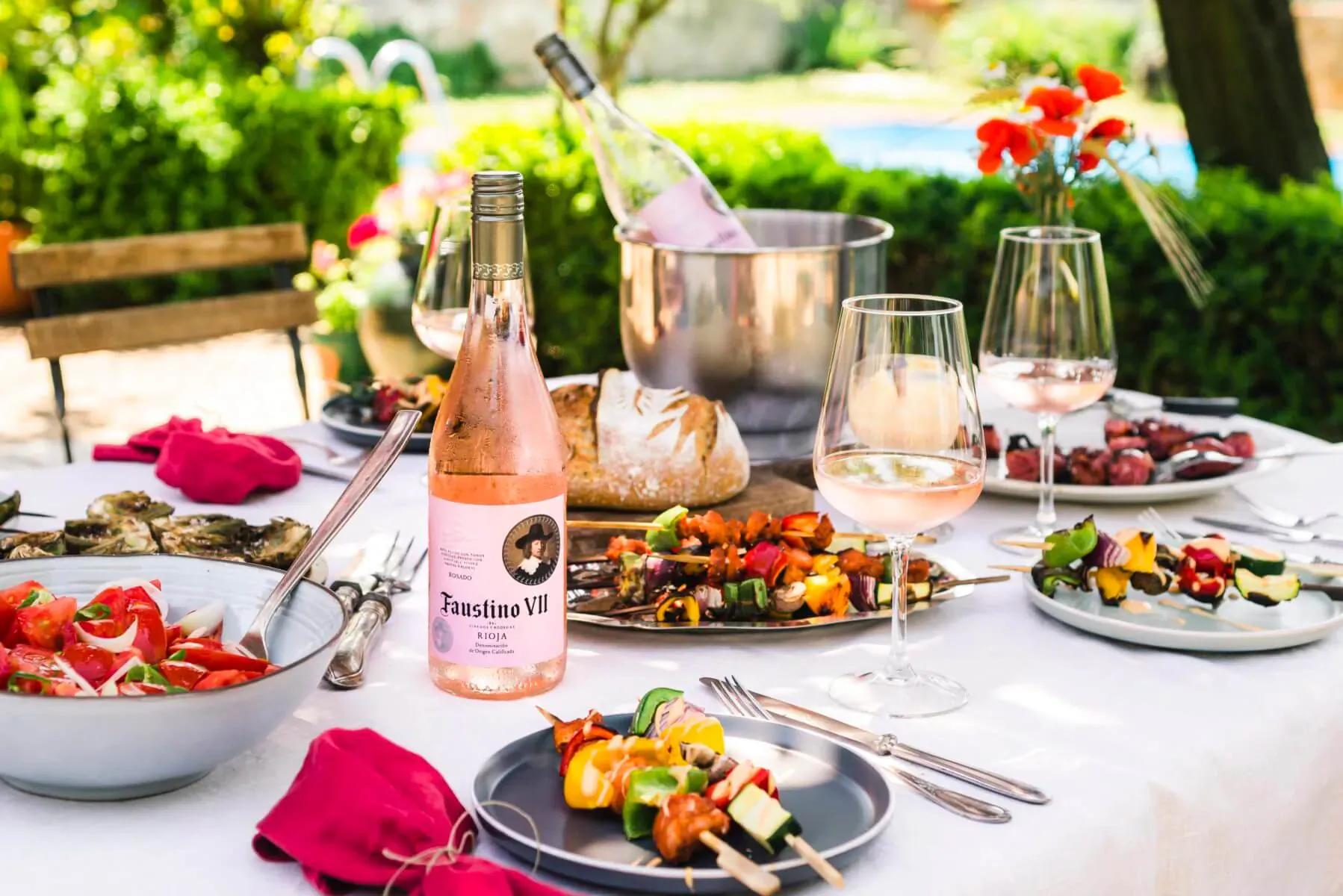 Outdoor table with chilled rosé wine bottle 'Faustino Rosé', bread, skewers, salads by poolside.