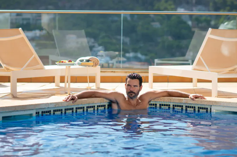 Shirtless man with arms outstretched relaxing in blue pool at luxury resort with lounge chairs and ocean view