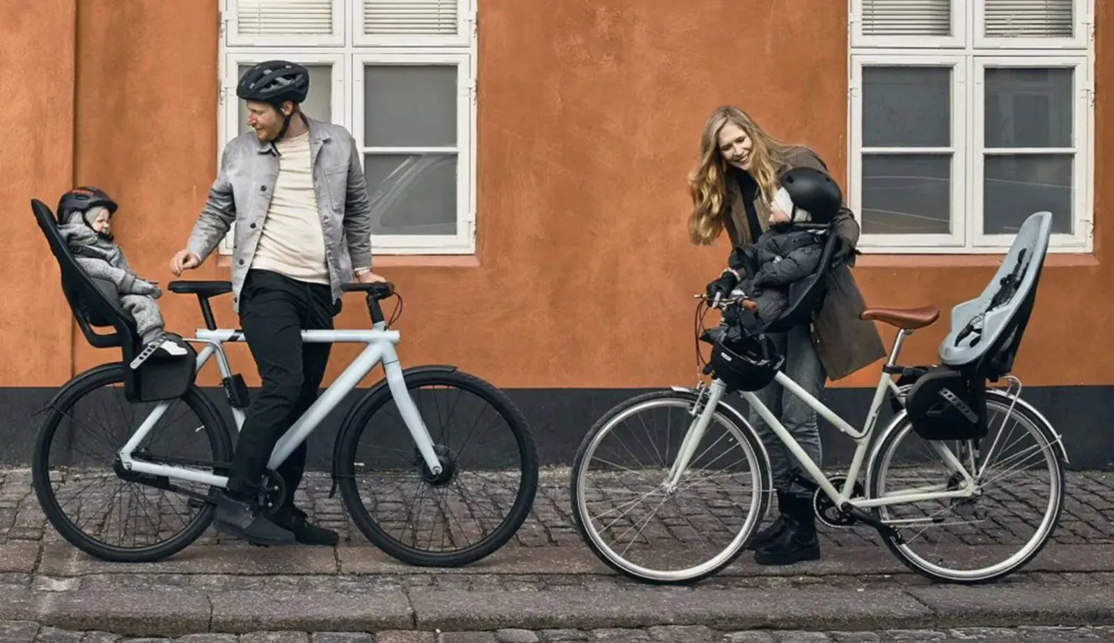 Family with dad in helmet and toddler in Thule Yepp 2 Mini front seat on white bike, mom with baby on another bike by orange wall.