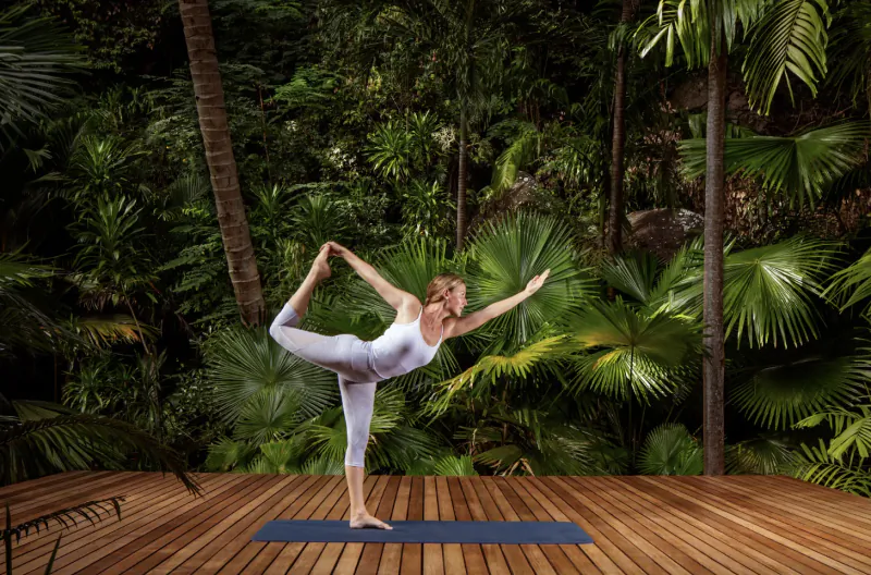 Blonde woman in white yoga outfit performs dancer pose on blue mat on wooden deck amid lush Seychelles palm trees