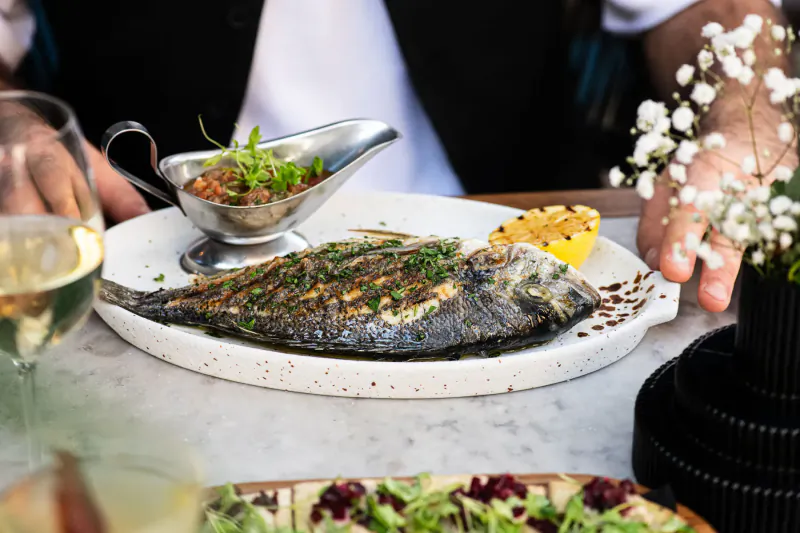 Grilled sea bream on white plate with lemon wedge, herb sauce, white wine glass, and garnishes at Mediterranean restaurant table