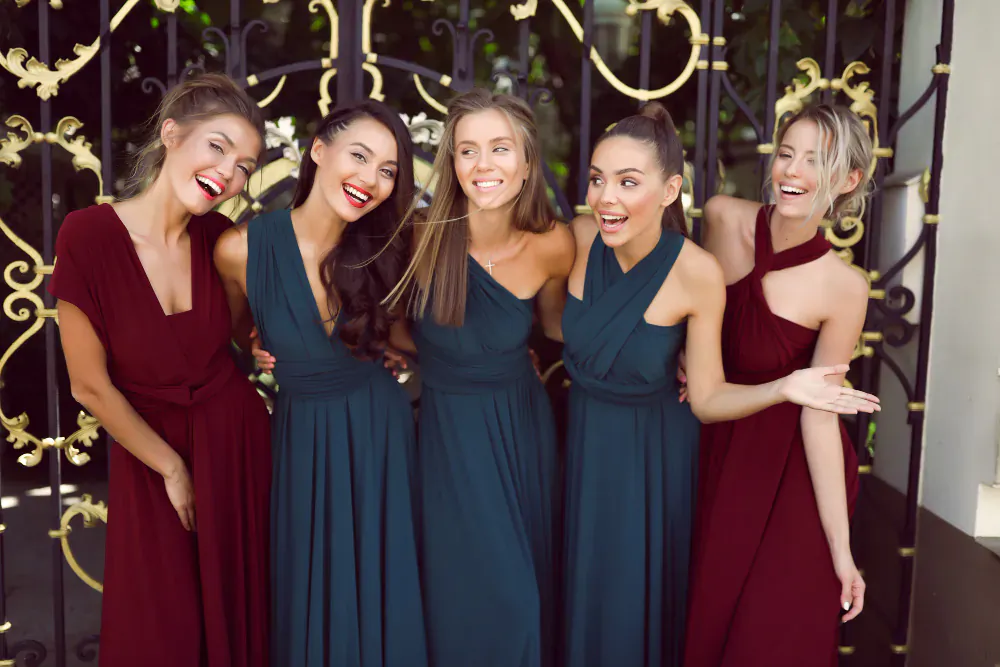 Five smiling women in elegant wrap dresses (emerald and burgundy) posing in front of ornate gold gate.