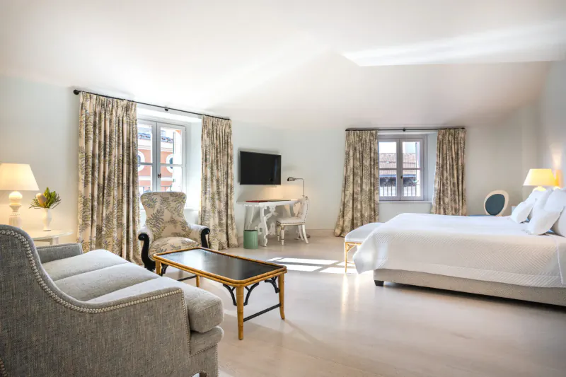 Elegant hotel suite with white bed, gray sofa, armchairs, desk, TV, and large curtained windows