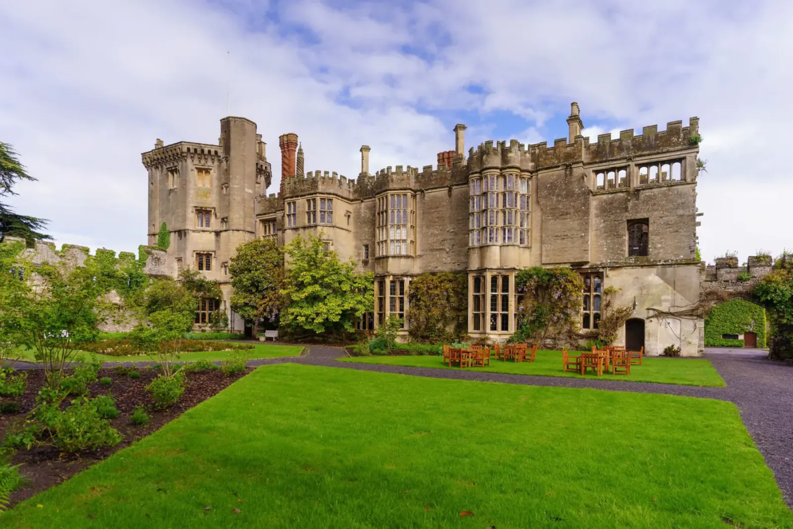 Thornbury Castle, a historic turreted manor with gardens, lawn, and benches under cloudy sky