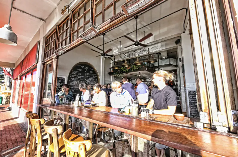 Customers at wooden bar counter in Kalk Bay cafe, chatting over drinks, open windows to street, pendant lights.