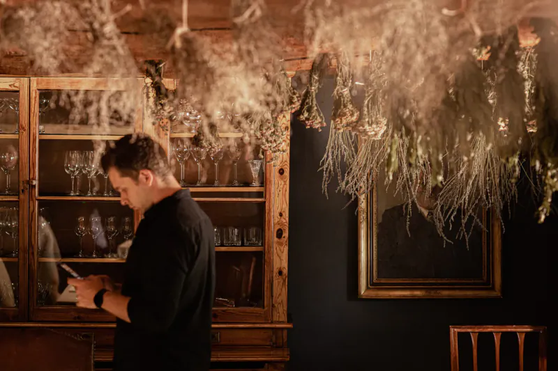 Man in black shirt reads paper near wooden cabinet with glassware and hanging herbs in cozy dining room