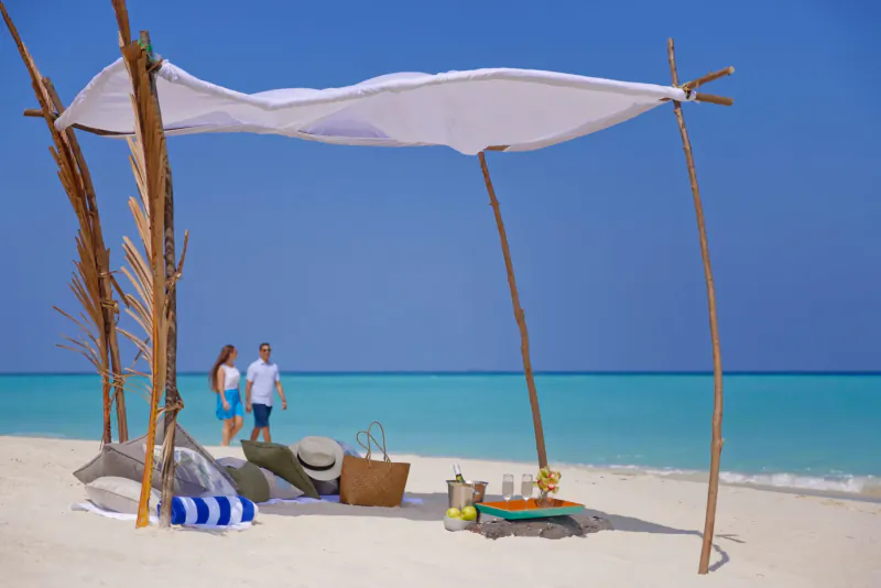 Couple in white and blue near shaded beach setup with drinks, basket, towels on white sand by turquoise sea, Maldives