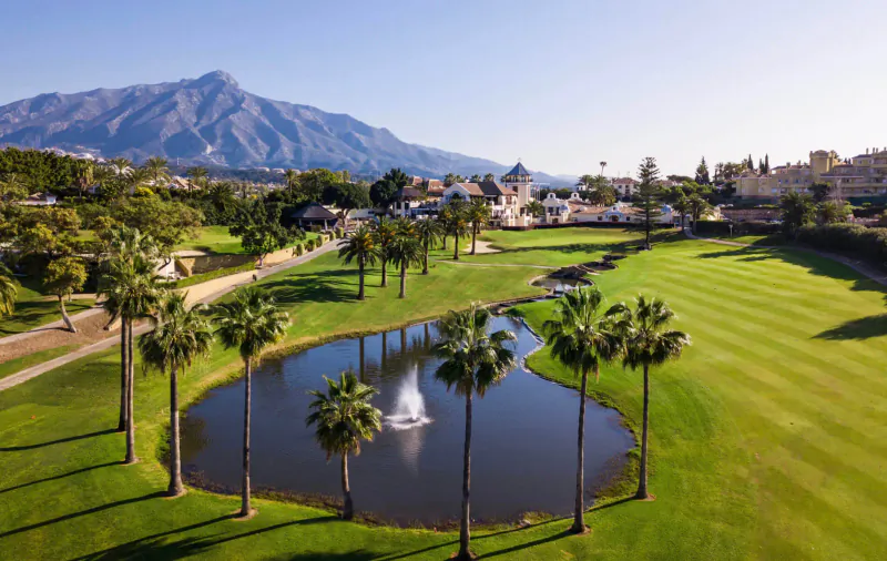 Aerial view of Los Naranjos Golf Club in Marbella: lush green fairway, palm-fringed pond with fountain, Sierra Blanca mountains backdrop.