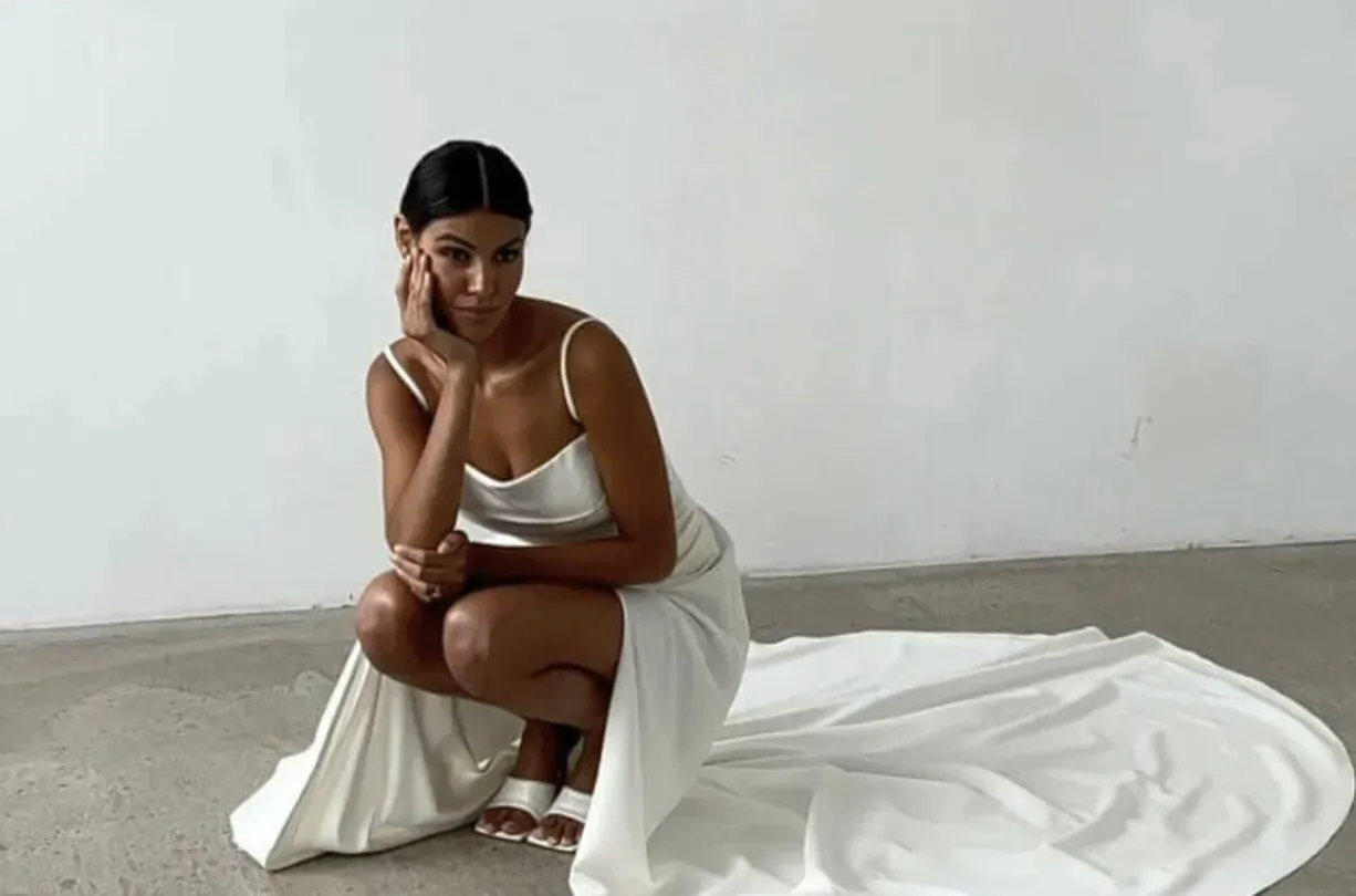 Brunette bride in white slip gown with train kneels on studio floor, hand on cheek