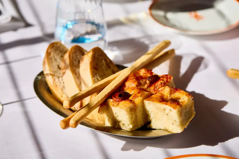 Oval dish of golden focaccia topped with tomatoes, garnished with grissini breadsticks, beside water glass on table at Italian restaurant.